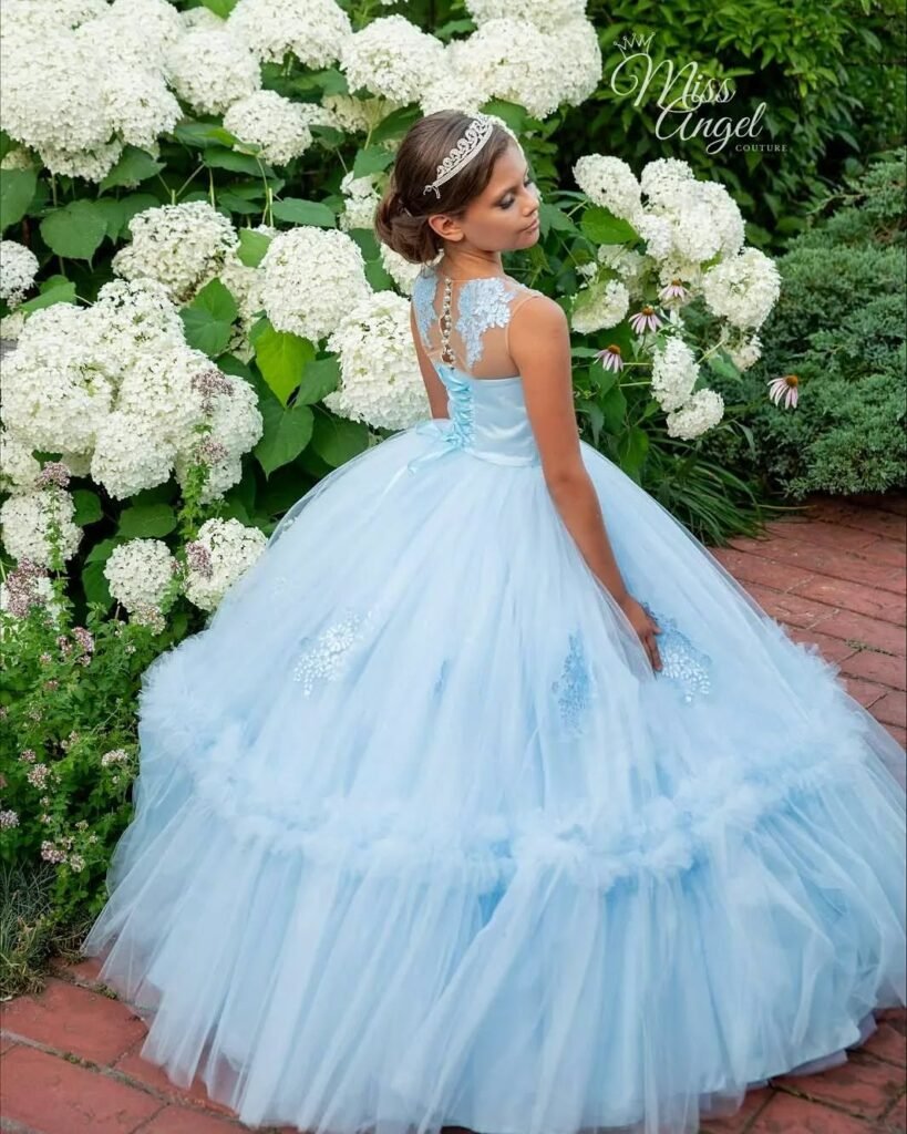Girl wearing an elegant light blue ball gown with lace and tulle, standing in a garden of white hydrangeas – Miss Angel Couture luxury fashion