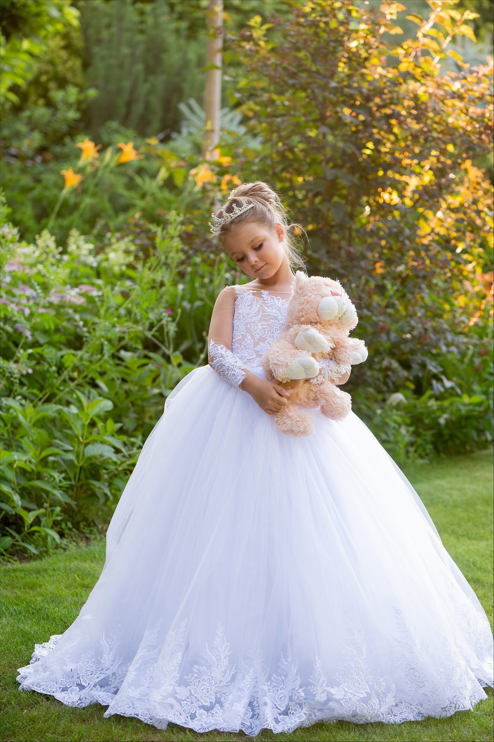 Elegant young girl in white lace ball gown holding teddy bear in sunlit garden, evoking fairy-tale charm and special occasion style.