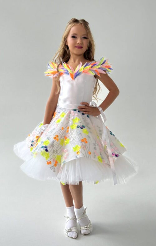 Young girl in a white satin and tulle dress with multicolored floral appliqués and feathered shoulder details, standing in a festive outdoor setting