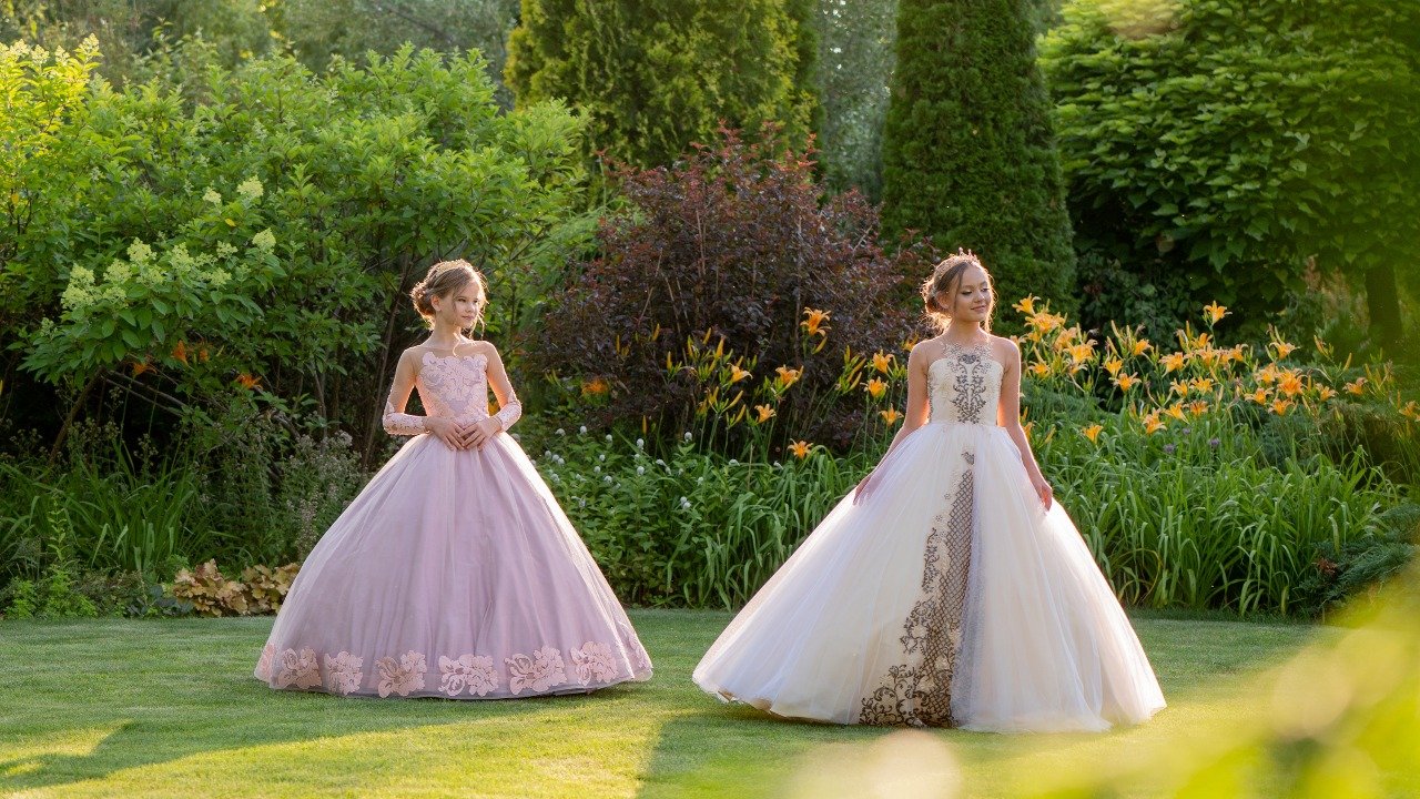 Two girls wearing elegant celebration ball gowns in a vibrant garden during golden hour—one in a pink dress, the other in white—posing for Miss Angel Couture’s luxury fashion campaign.