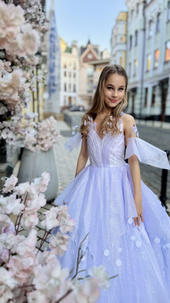 Girl in a lavender tulle ball gown with off-shoulder floral sleeves, standing on a cobblestone street with pastel buildings and flower arrangements