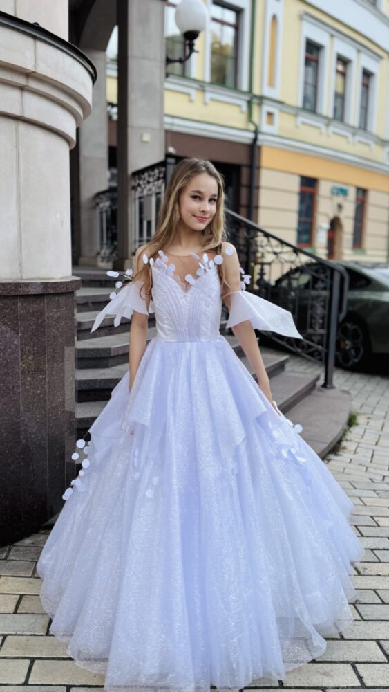 Young girl wearing a floor-length ivory tulle gown with floral appliqués and sheer off-shoulder sleeves, standing outdoors near a staircase