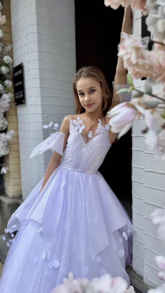 Young girl in a shimmering white tulle gown with petal appliqués and sweetheart neckline, standing in front of a floral-decorated white brick wall