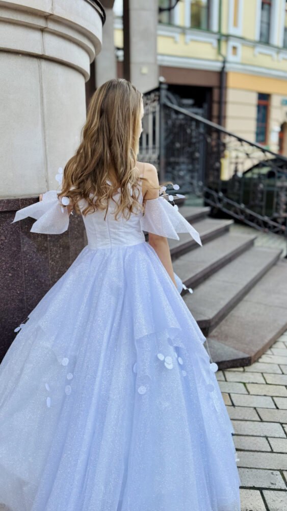 Girl with long wavy hair wearing a white floor-length tulle gown with sheer bow sleeves and floral appliqués, standing on stone steps near an ornate railing