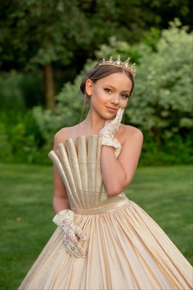 Grace gold gown with fan-pleated bodice, shimmering full skirt, jeweled crown, and lace gloves—styled outdoors in a lush garden for royal-themed events, weddings, and fantasy celebrations.