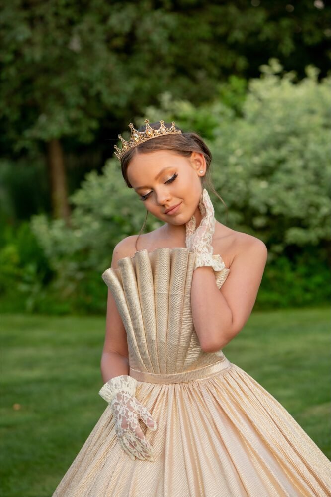 Grace gold gown with fan-pleated bodice, voluminous textured skirt, jeweled crown, and lace gloves—styled outdoors in a garden setting for royal-themed events, weddings, and pageants.