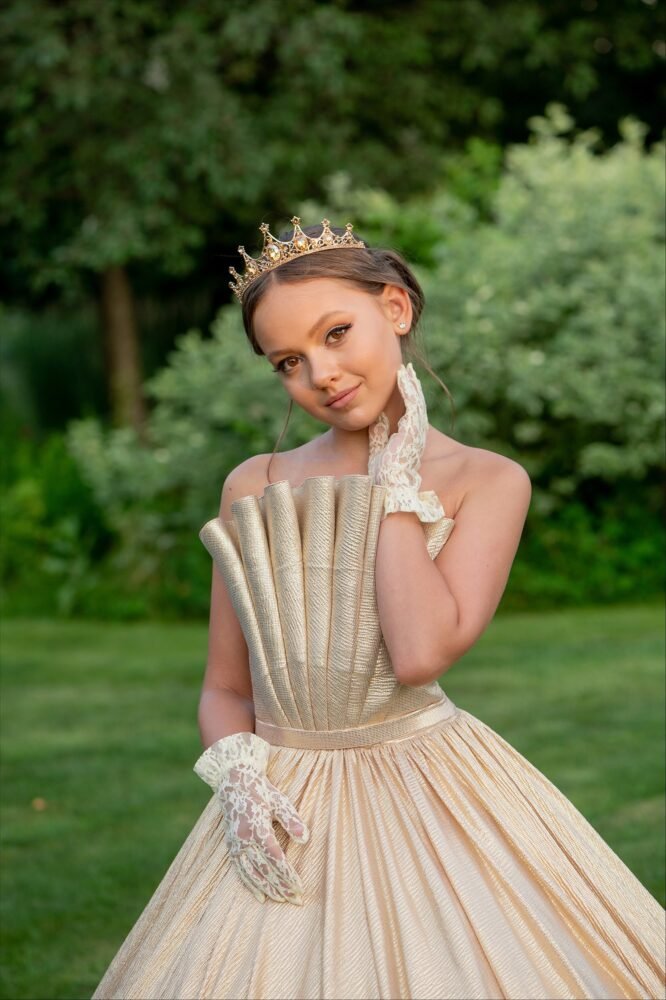 Grace gown for girls in shimmering gold with fan-pleated bodice, voluminous skirt, jeweled crown, and lace gloves—styled outdoors for weddings, pageants, and royal-themed celebrations.