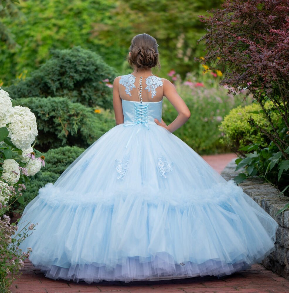 Back view of Isabella light blue ball gown with corset-style ribbon lacing, floral appliqués, and ruffled tulle hem—styled outdoors on a brick pathway with hydrangeas and garden greenery for weddings, fantasy shoots, and royal-themed celebrations.
