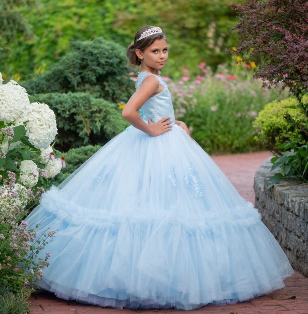 Isabella light blue ball gown with floral embellishments, layered tulle skirt, and silver tiara—styled outdoors on a brick pathway with blooming garden backdrop for weddings, fantasy shoots, and royal-themed celebrations.