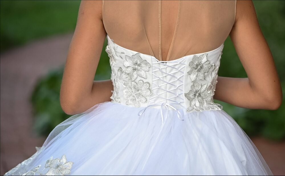 Back view of Layla white ball gown with lace-up corset, sheer shoulder panel, silver floral embroidery, and layered tulle skirt—styled outdoors on a brick pathway for weddings, fantasy shoots, and royal-themed celebrations.