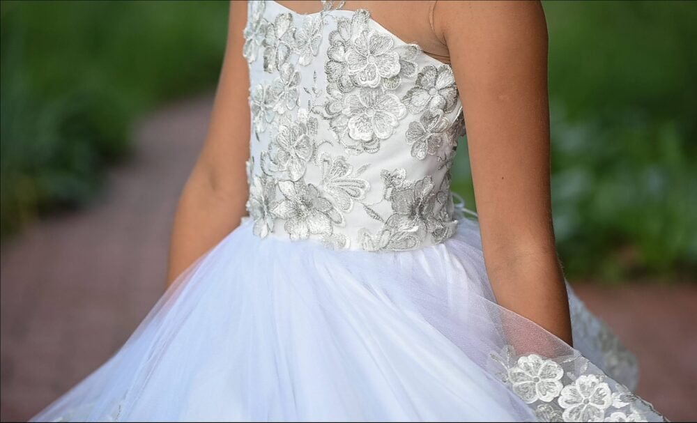Close-up of Layla white ball gown with silver floral embroidery and layered tulle skirt—styled outdoors against greenery and brick pathway for weddings, fantasy shoots, and royal-themed celebrations.