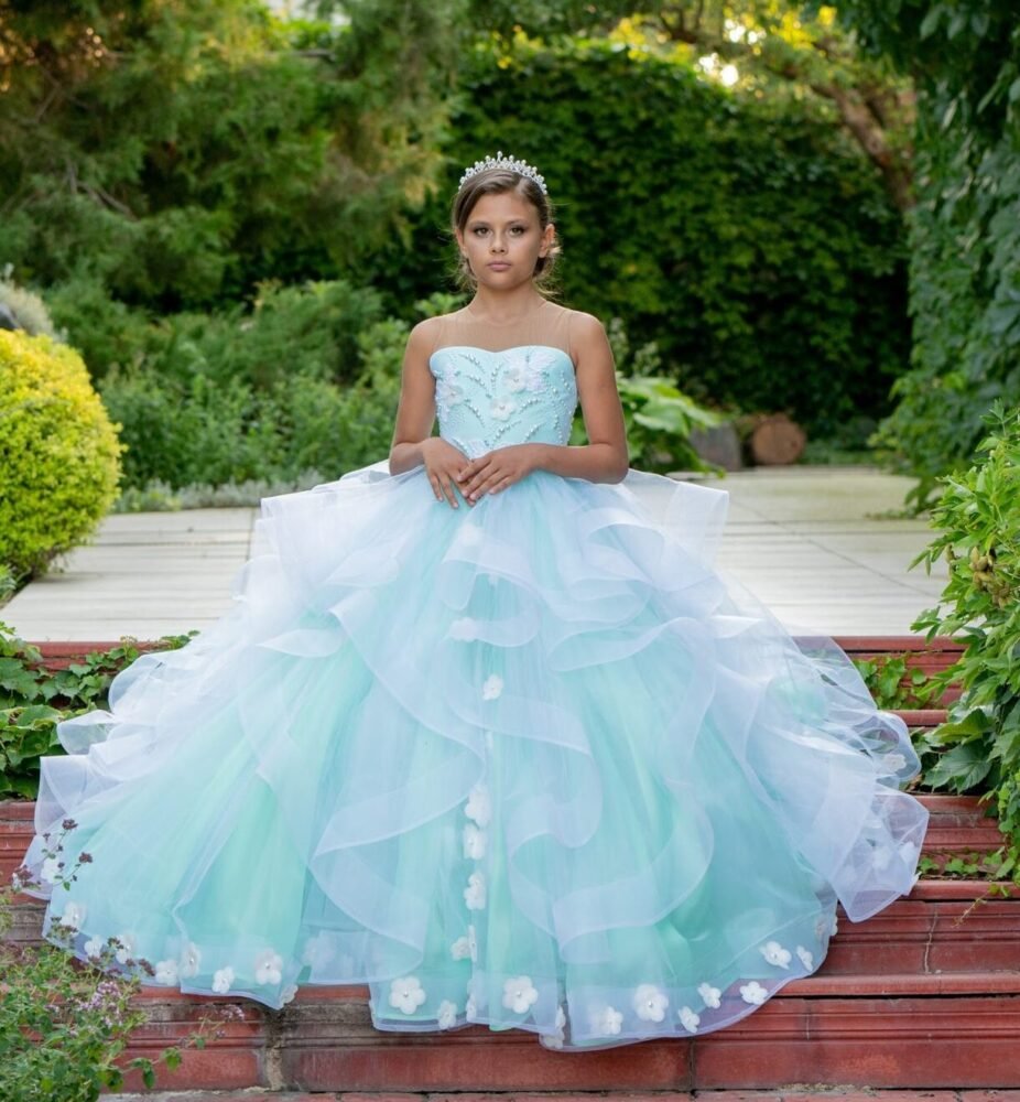 Olivia light blue ball gown with strapless sweetheart neckline, layered tulle skirt, white floral hem accents, and silver tiara—styled outdoors on a red-tiled pathway with lush greenery for weddings, fantasy shoots, and royal-themed celebrations.