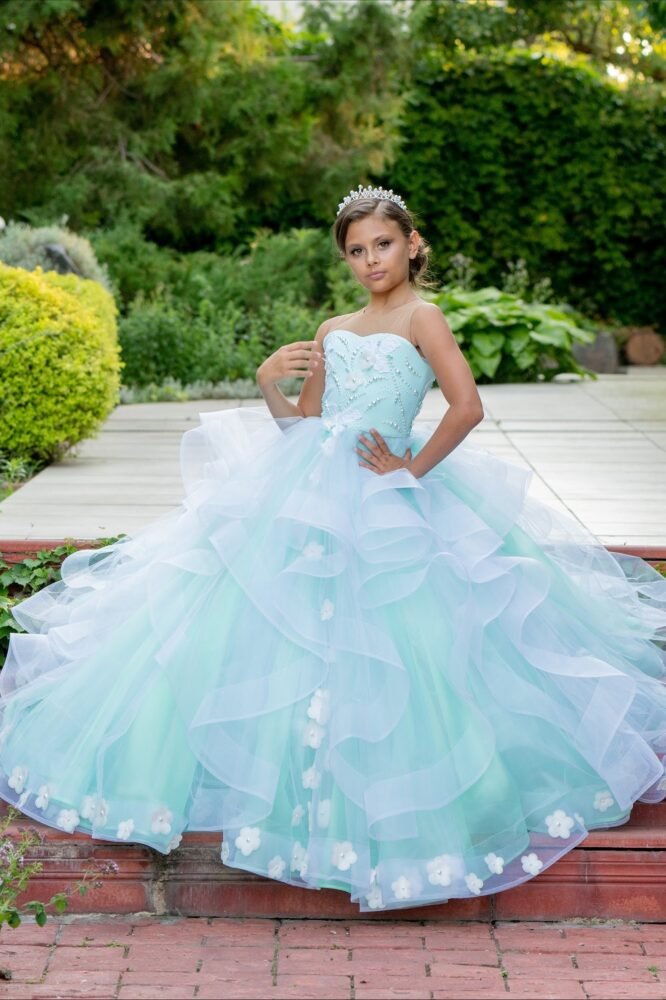 Olivia light blue ball gown with layered tulle skirt, floral hem embellishments, and sequin bodice—styled outdoors in a garden setting with tiara for weddings, fantasy shoots, and royal-themed celebrations.