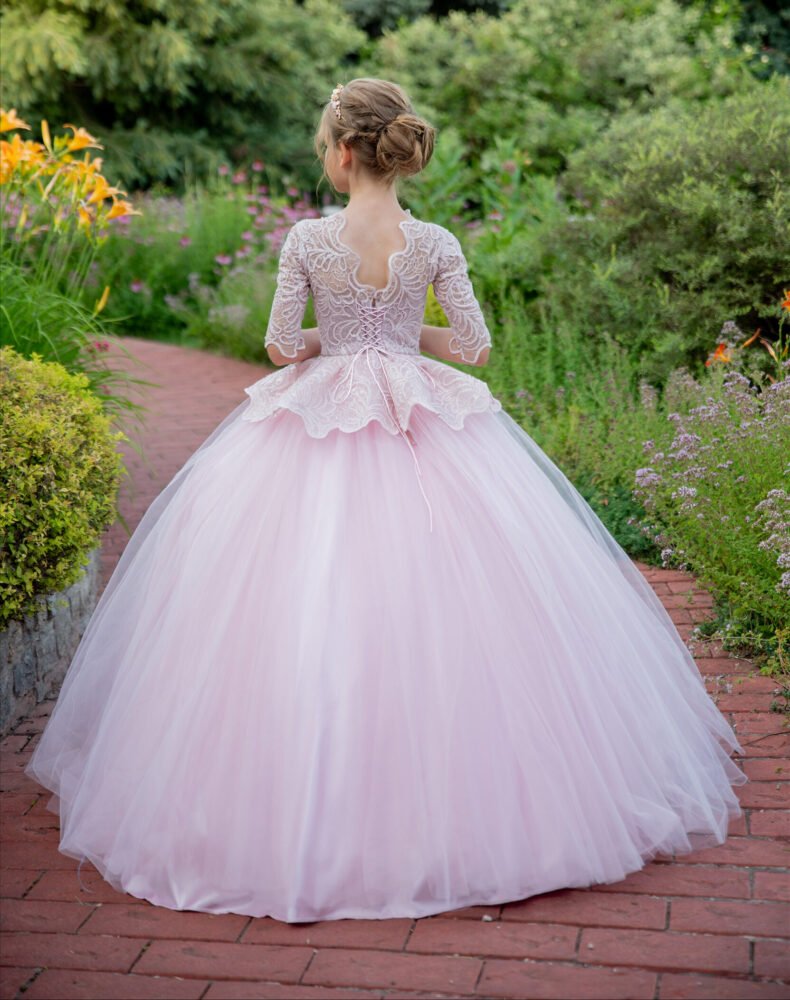 Back view of Rosie light pink ball gown with scalloped lace bodice, three-quarter sleeves, and layered tulle skirt—styled outdoors on a brick pathway with blooming lilies for weddings, fantasy shoots, and royal-themed celebrations.