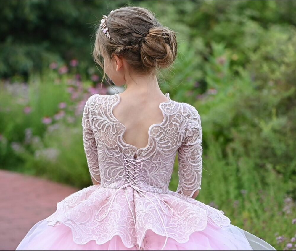 Back view of Rosie light pink ball gown with scalloped lace overlay, corset-style lace-up back, and voluminous tulle skirt—styled outdoors in a garden with pink flowers for weddings, fantasy shoots, and royal-themed celebrations.