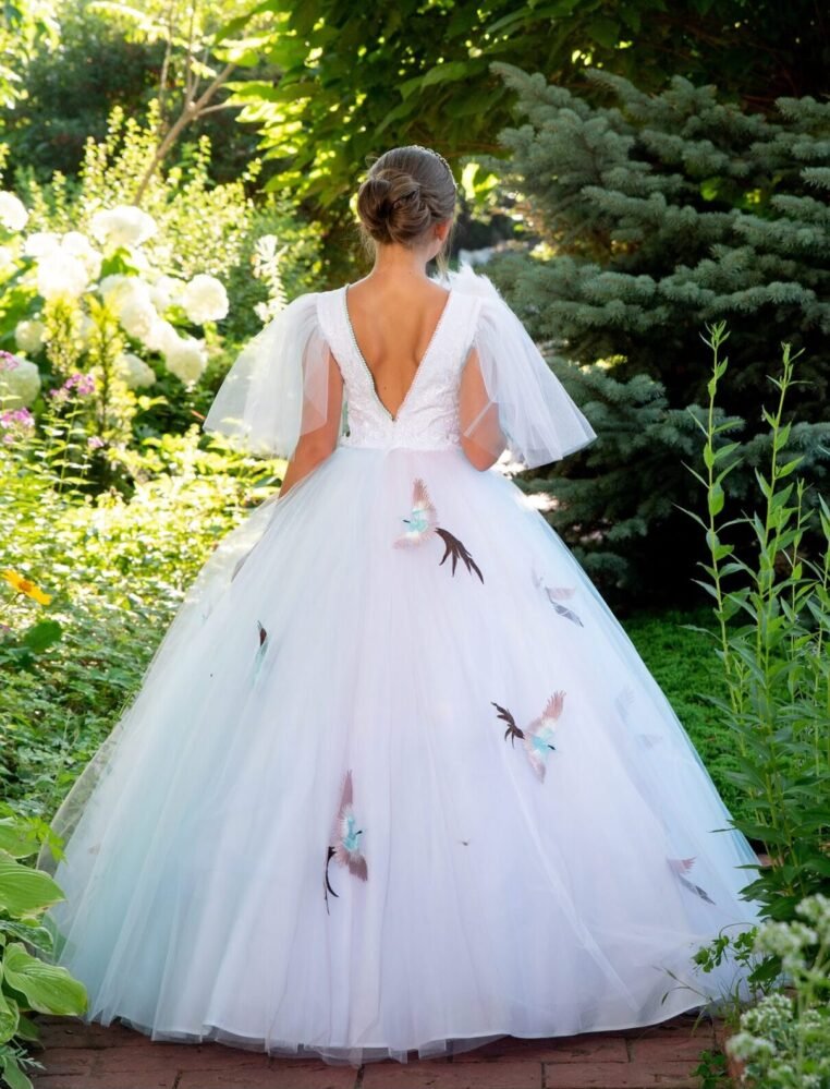 Back view of Sophia white ball gown with deep V-back, sheer puff sleeves, and bird appliqués on layered tulle skirt—styled outdoors on a brick path with garden greenery and white flowers for weddings, fantasy shoots, and royal-themed celebrations.