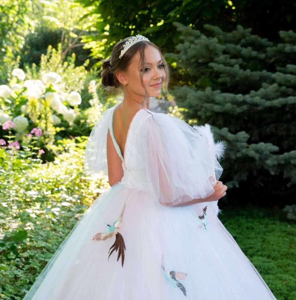 Back view of Sophia white ball gown with deep V-back, sheer puff sleeves, and bird embroidery on layered tulle skirt—styled outdoors in a garden with tiara and flowering greenery for weddings, fantasy shoots, and royal-themed celebrations.