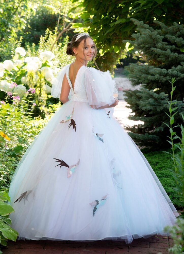 Sophia white ball gown with deep V-back, puff sleeves, and bird appliqués on layered tulle skirt—styled outdoors in a garden with tiara and smiling pose for weddings, fantasy shoots, and royal-themed celebrations.
