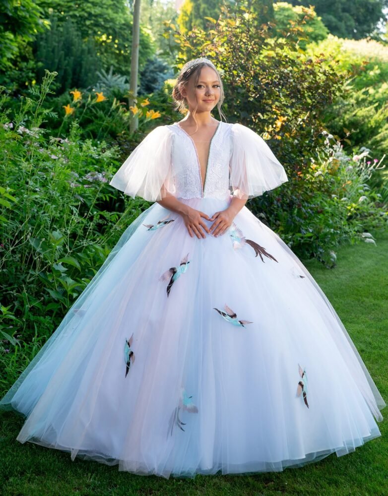 Sophia white ball gown with deep V-neckline, voluminous tulle sleeves, and colorful bird appliqués—styled outdoors in a sunlit garden with floral headpiece for weddings, fantasy shoots, and royal-themed celebrations.