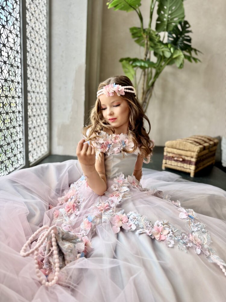 Girl seated indoors wearing ivory ball gown with pastel floral appliqués, pearl headband, and matching floral handbag